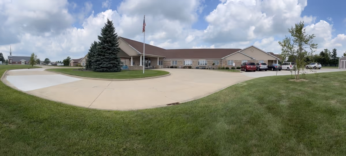 Wide exterior view of a single-story senior living facility building with a brown roof and beige walls, surrounded by a large concrete driveway and green grass. There is a tall evergreen tree and an American flag on a flagpole in front of the building. Several cars are parked near the entrance under a partly cloudy sky.