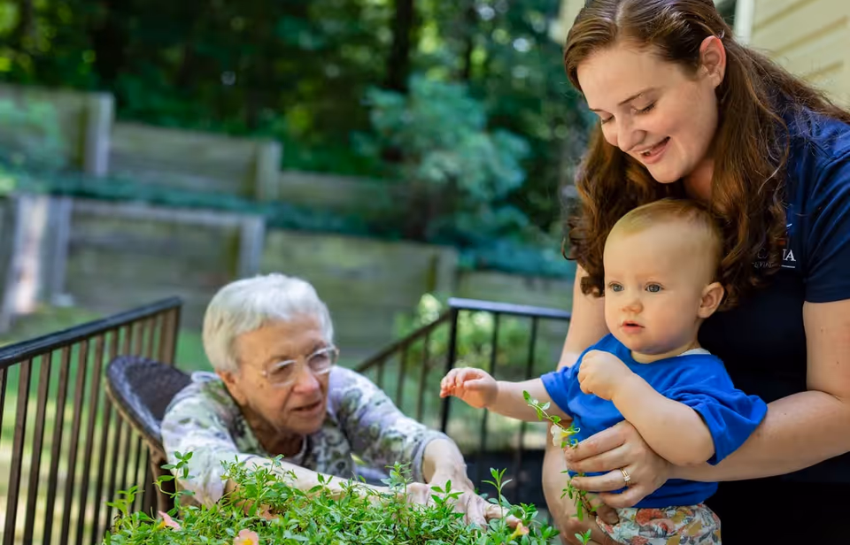 An elderly woman sitting outdoors near a garden bed, reaching towards some plants, while a young woman holding a baby watches and smiles. The background shows greenery and wooden retaining walls.