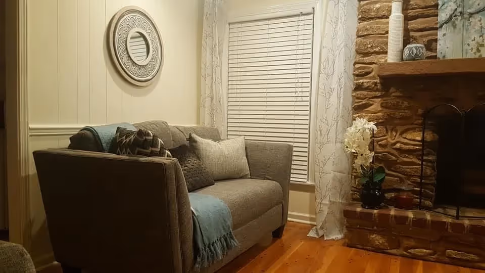 Cozy living room with a gray upholstered sofa adorned with multiple cushions and a blue throw blanket. A round decorative mirror hangs on the wall above the sofa. Next to the sofa is a window with white blinds and sheer curtains featuring a subtle branch pattern. To the right, there is a stone fireplace with a wooden mantel decorated with vases and a small plant.