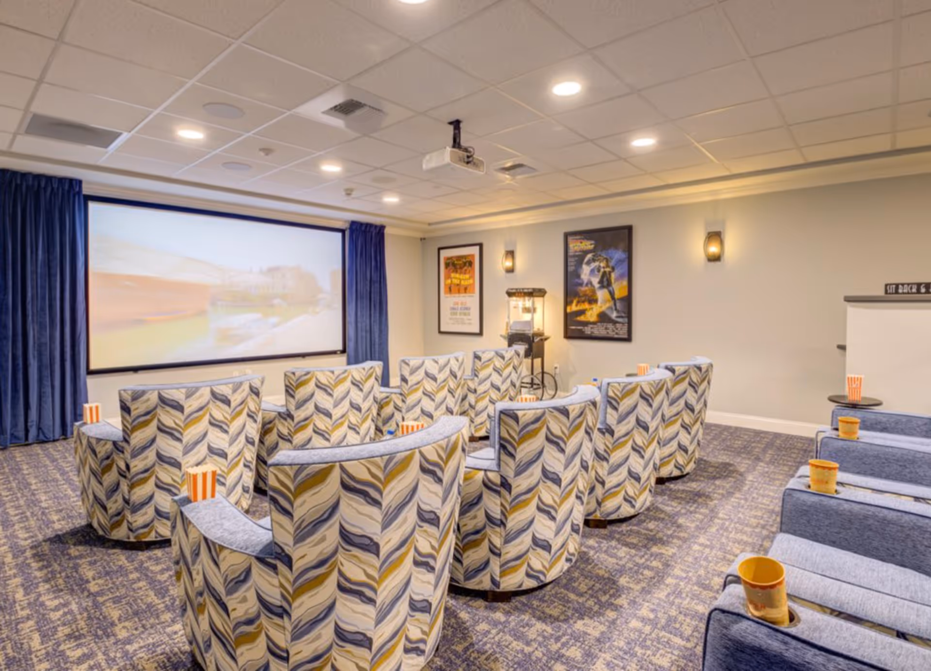 A cozy movie theater room with patterned armchairs arranged in rows facing a large projection screen. The room has a ceiling-mounted projector, blue curtains framing the screen, and movie posters on the walls. Popcorn containers are placed on the armrests of the chairs.
