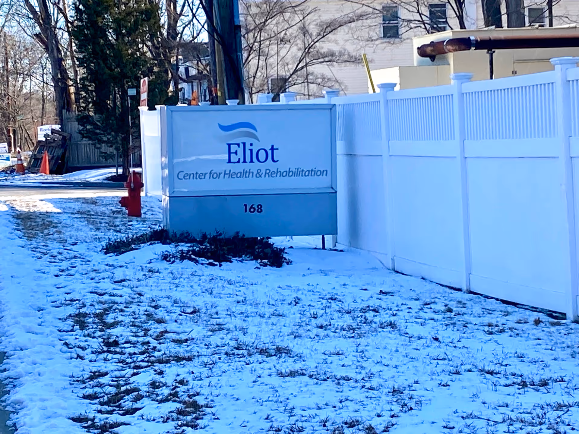 Outdoor view of a sign for Eliot Center for Health & Rehabilitation next to a white fence with snow on the ground and trees in the background.