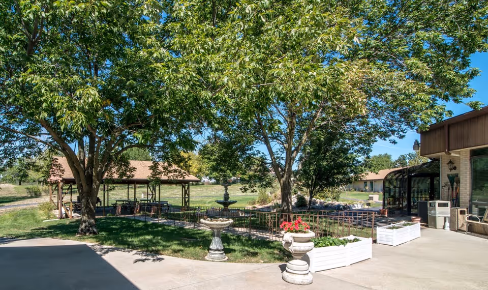 Outdoor patio area at SummitView Terrace featuring large leafy trees, a covered pavilion with seating, a decorative fountain, and several white planter boxes with flowers. The scene is sunny with a clear blue sky.