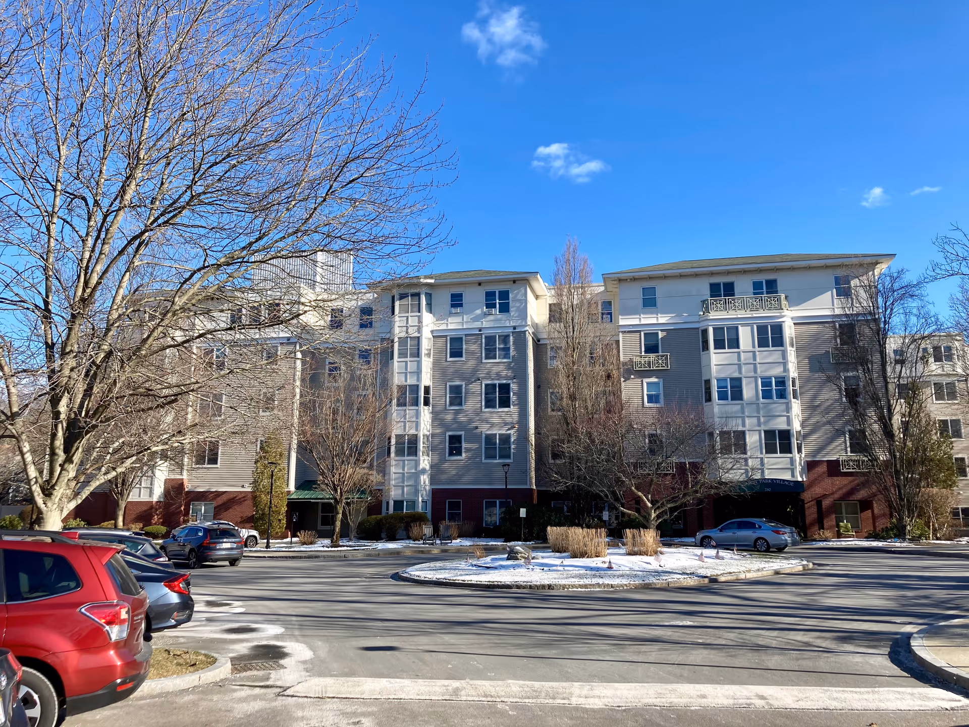 Front exterior of a multi-story senior living building with a circular driveway, parked cars, and leafless trees under a clear blue sky.