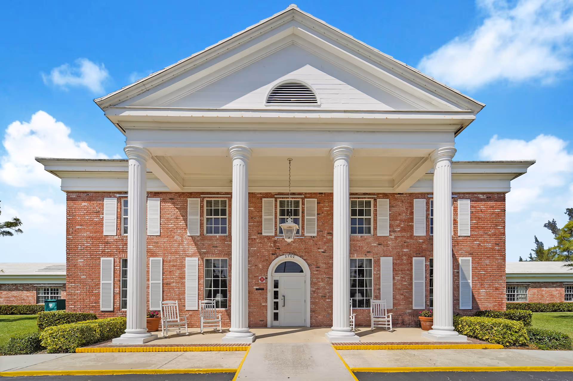 Front exterior view of a red brick building with white columns and a triangular pediment, featuring a covered entrance with rocking chairs on the porch and a clear blue sky in the background.
