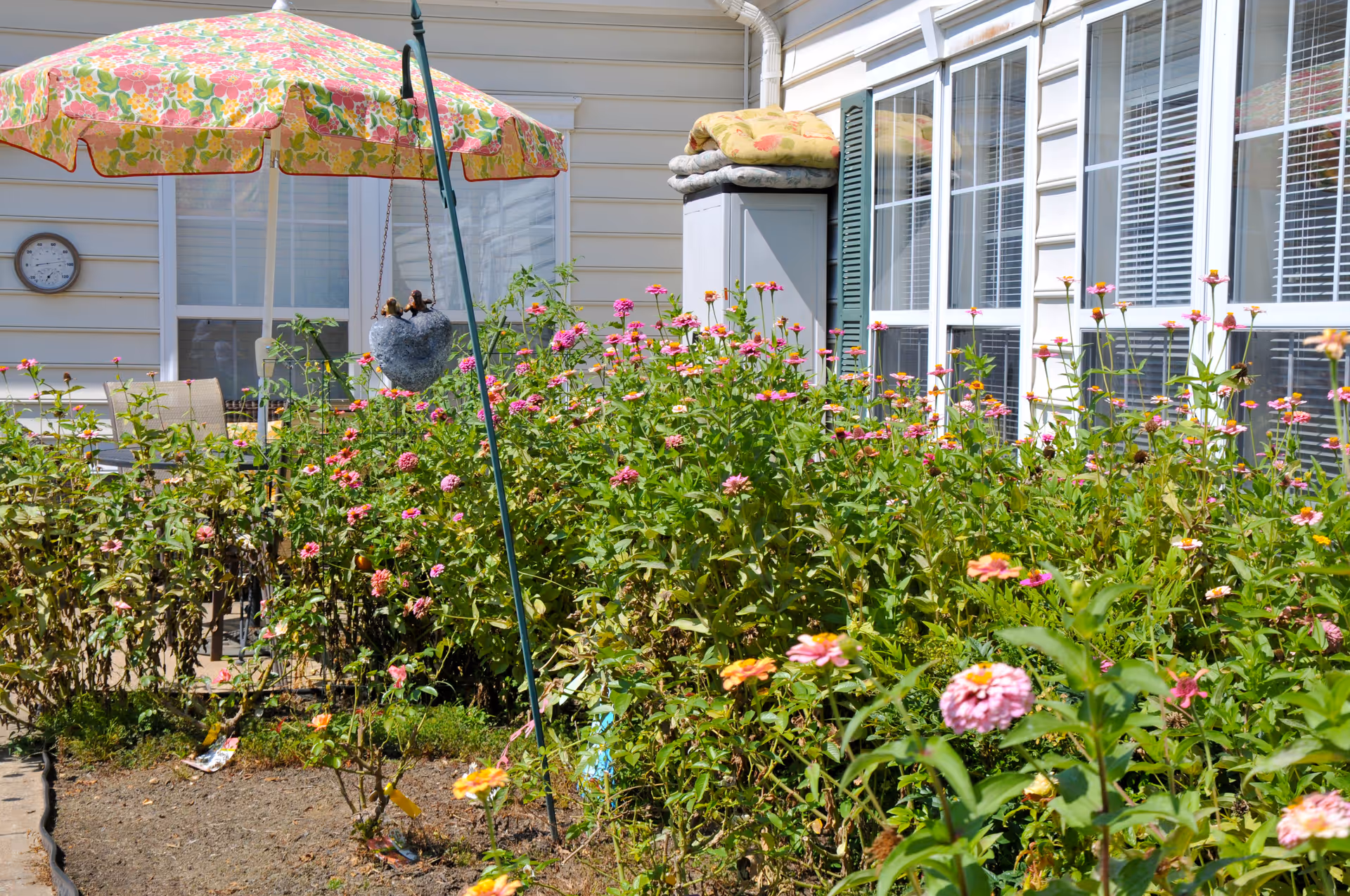 A garden area with blooming pink and orange flowers in front of a building with large windows. There is a colorful floral umbrella providing shade over a patio area with chairs, and a hanging planter with small plants. A storage cabinet with folded cushions on top is also visible near the windows.