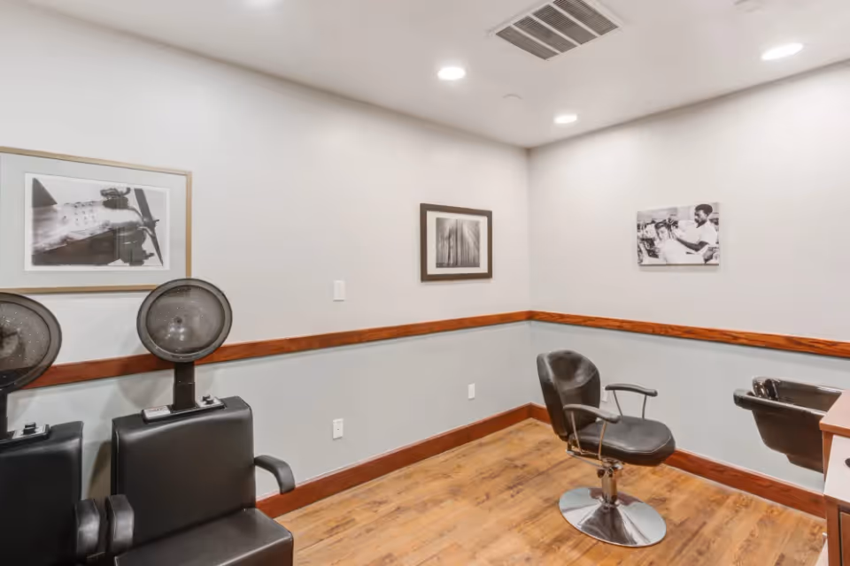 Interior view of a hair salon area in a senior living facility with two black hair drying chairs on the left and a black styling chair in front of a hair washing sink on the right. The room has light gray walls with a wooden chair rail, wood flooring, and three framed black and white pictures on the walls.