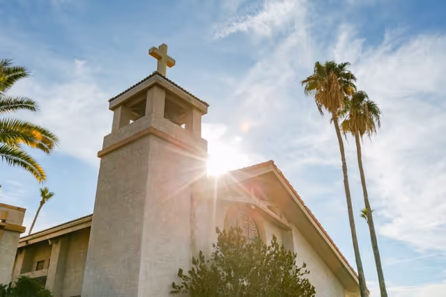 Sunlit church-style building with a bell tower topped by a cross and tall palm trees against a blue sky.
