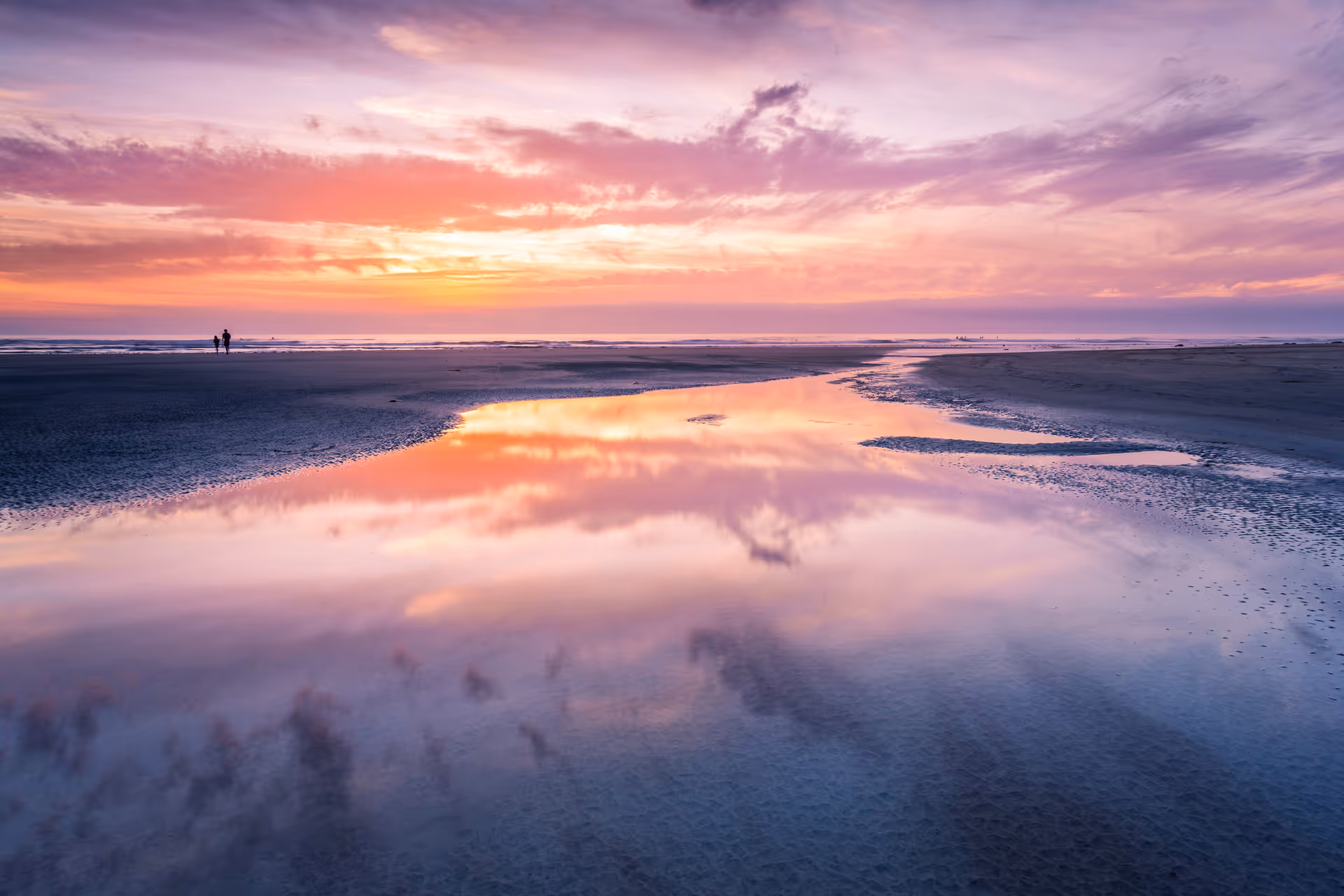 A serene beach scene at sunset with a colorful sky reflecting on a shallow water pool on the sand. Two people are walking along the shoreline in the distance.