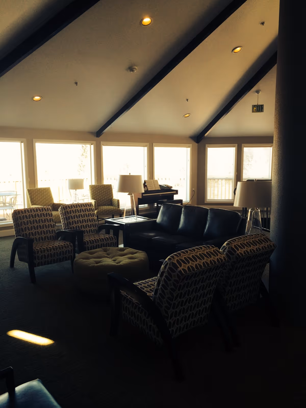 A cozy living room area with patterned armchairs, a black leather sofa, a round tufted ottoman, and several table lamps. Large windows let in natural light, and a piano is visible in the background. The ceiling has exposed beams with recessed lighting.