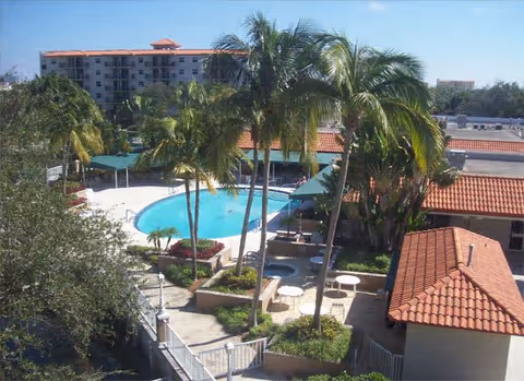 Outdoor view of a senior living facility courtyard featuring a swimming pool surrounded by palm trees, patio tables with umbrellas, and buildings with red-tiled roofs under a clear blue sky.