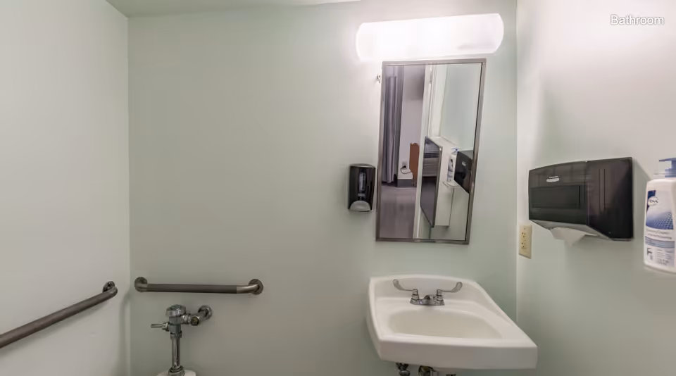 A small bathroom with a white sink, a mirror above it, a wall-mounted soap dispenser, a paper towel dispenser, and grab bars on the walls. The walls are painted light green, and the lighting fixture above the mirror is turned on.