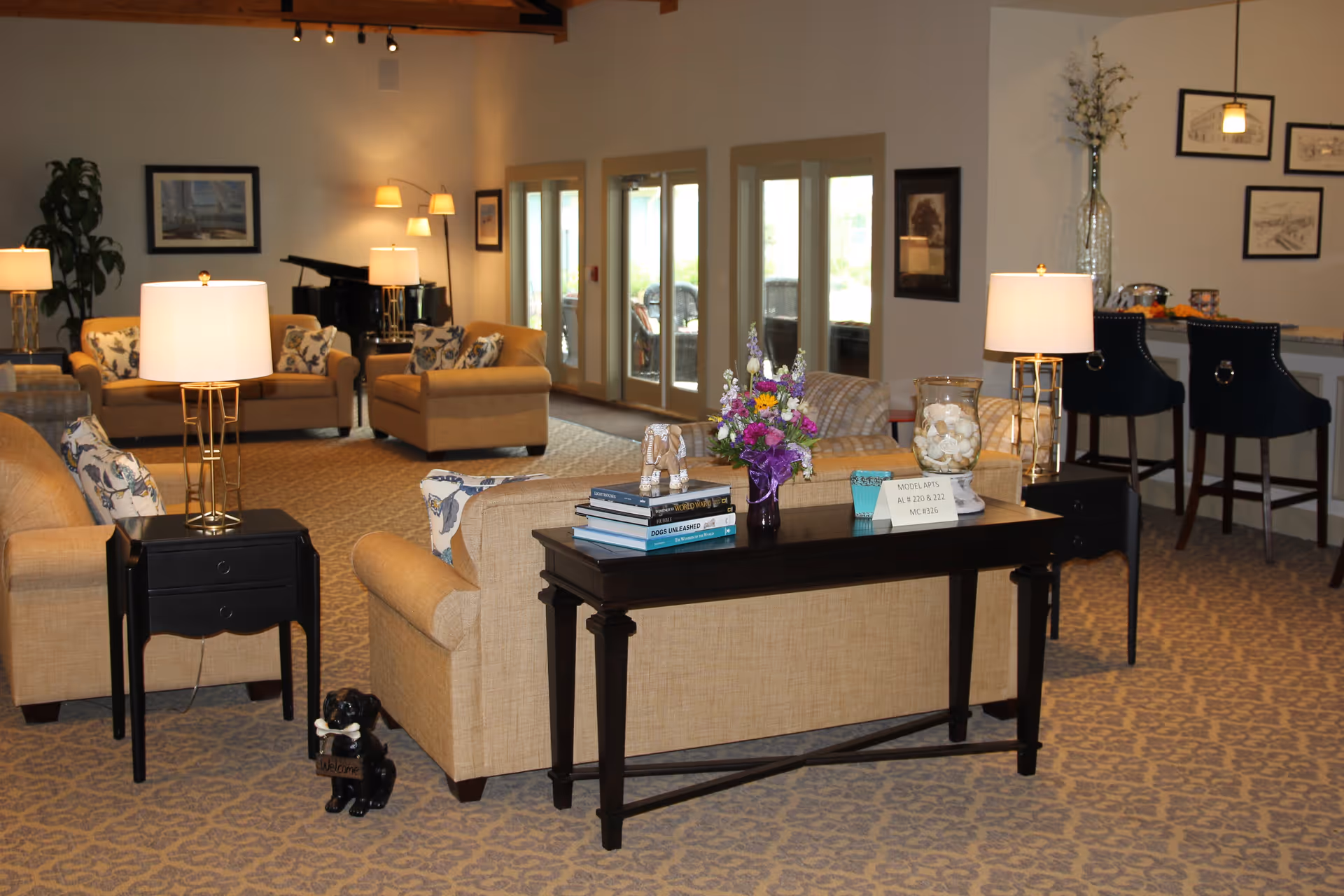 Communal living room with beige sofas, side tables and lamps, a console table with flowers and books, and a piano in the background.