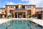 Outdoor view of La Posada at Green Valley featuring a large swimming pool in the foreground with clear water, surrounded by poolside chairs and tables. The building has a warm beige and terracotta color scheme with multiple windows, balconies, and a covered entrance area under a bright blue sky with some clouds.