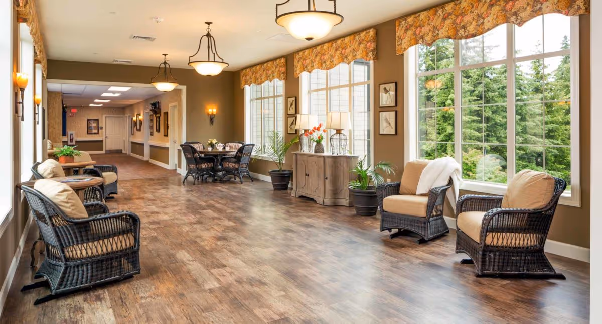A spacious, well-lit sitting area in a senior living facility with large windows showing green trees outside. The room features wicker chairs with cushions, small tables, potted plants, a wooden sideboard with lamps and flowers, and a dining table with chairs in the background. Warm lighting fixtures hang from the ceiling and wall sconces add additional light.