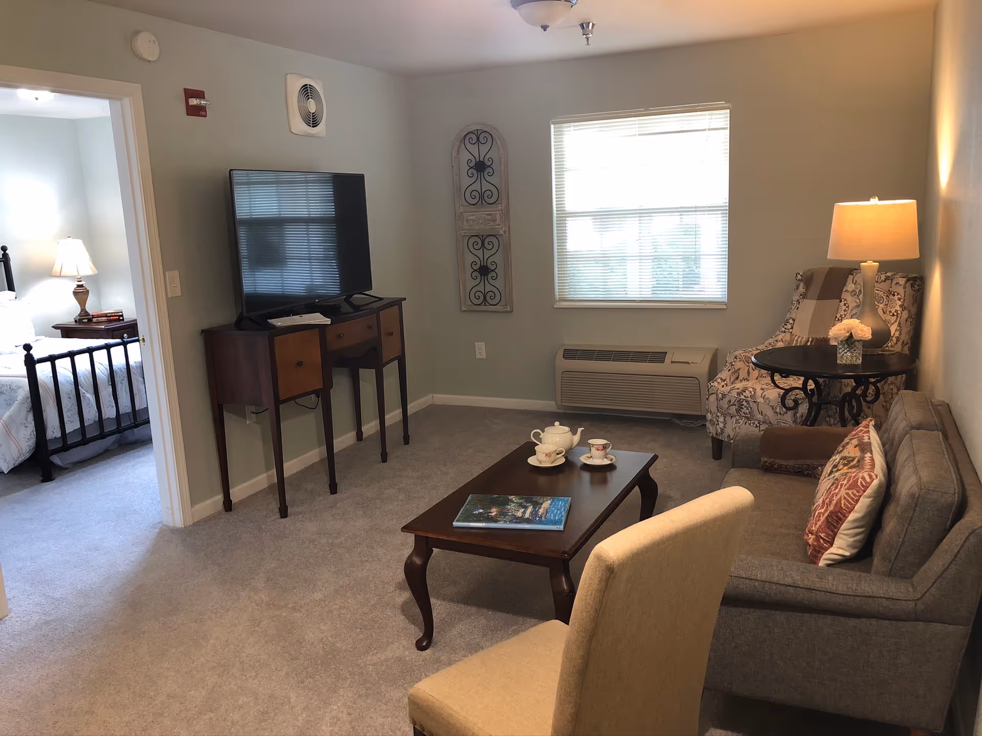 A cozy living room in an assisted living facility with a gray sofa, a patterned armchair with a lamp on a side table, a wooden coffee table with a tea set and a book, and a TV on a wooden stand. A window with blinds lets in natural light. An adjacent bedroom with a bed and a nightstand with a lamp is visible through an open doorway.
