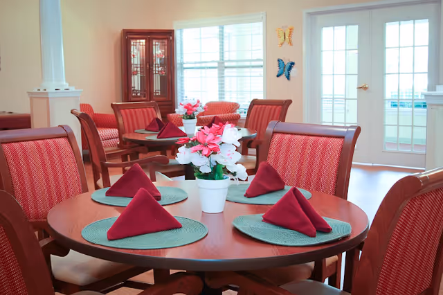 A bright dining room with round wooden tables set with green placemats and folded burgundy napkins. Each table has a white pot with pink and white flowers. The room has red cushioned chairs, a wooden cabinet with glass doors, large windows, and glass double doors leading outside. Butterfly wall decorations are visible near the doors.