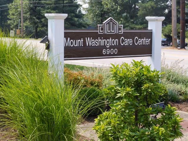 A wooden sign for Mount Washington Care Center with the number 6900, surrounded by green bushes and tall grass, located near a road with trees in the background.