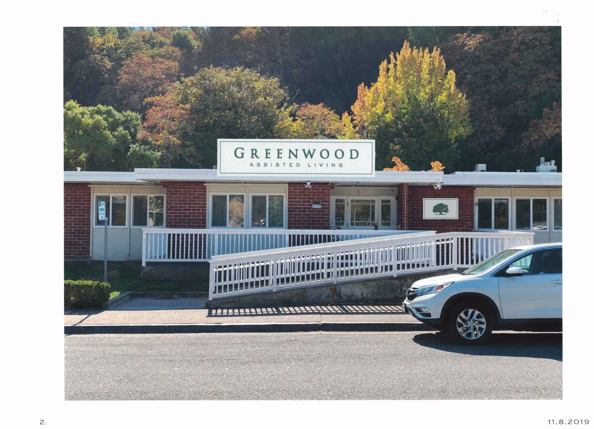 Front exterior view of Greenwood Assisted Living facility with a white ramp leading to the entrance, a white car parked on the street in front, and trees with autumn foliage in the background.