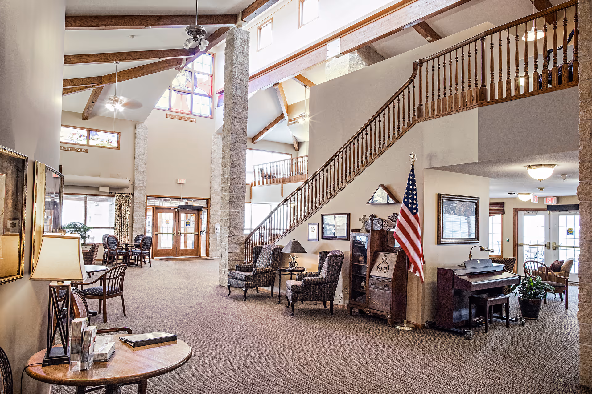 Spacious senior living facility common area with high ceilings, exposed wooden beams, and large windows allowing natural light. The room features comfortable seating including armchairs and small tables, a wooden piano with a bench, an American flag, and decorative framed pictures on the walls. A staircase with wooden railings leads to an upper level. The entrance doors are visible in the background.