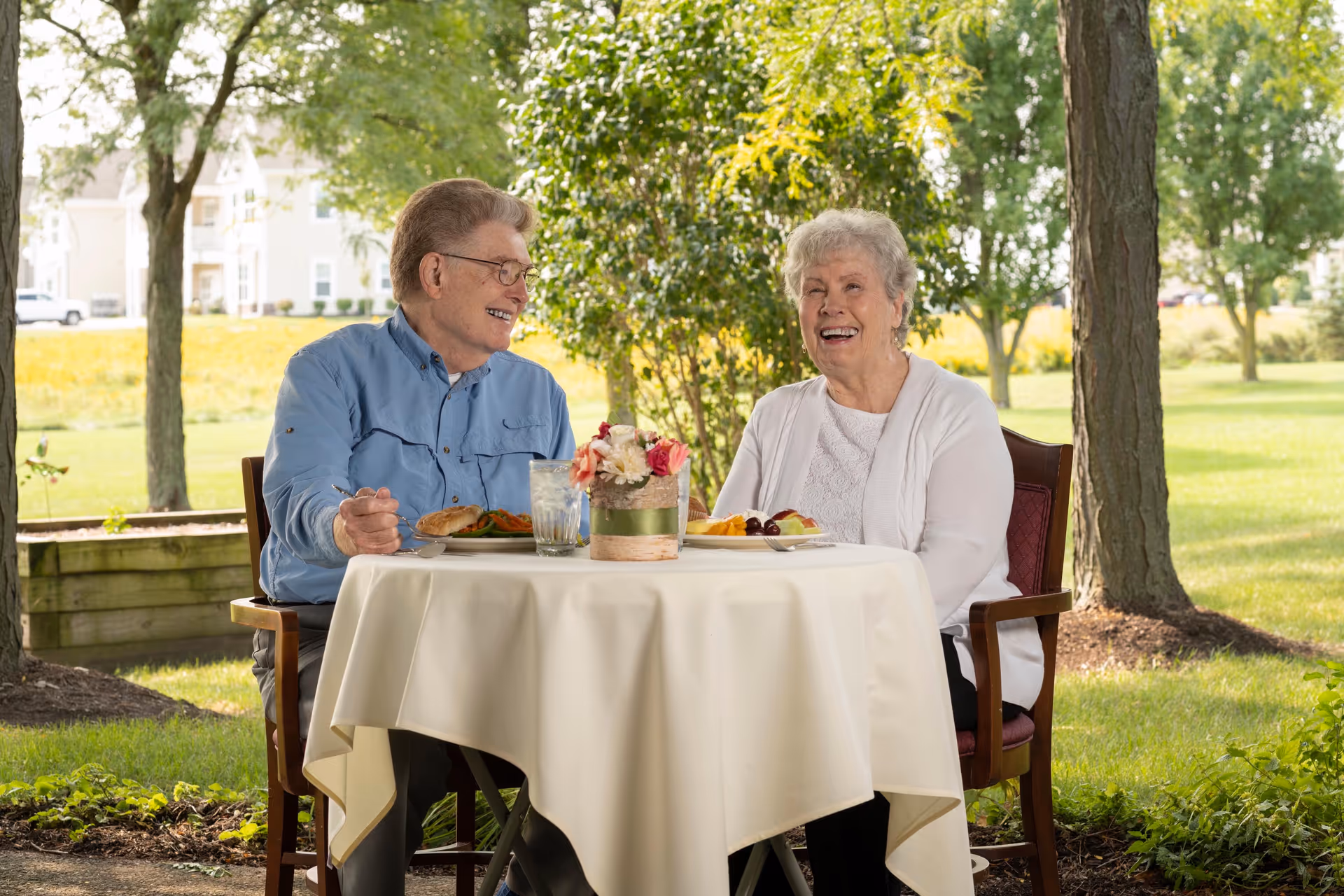 An elderly man and woman sitting at a round table covered with a white tablecloth outdoors, enjoying a meal together. The man is wearing a blue shirt and glasses, and the woman is wearing a white cardigan. They are smiling and appear to be having a pleasant conversation. The background shows green trees and a grassy area in a sunny setting.