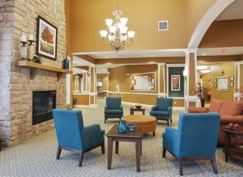Open seating lounge in an assisted living facility with teal armchairs and a round coffee table facing a stone fireplace under a chandelier.