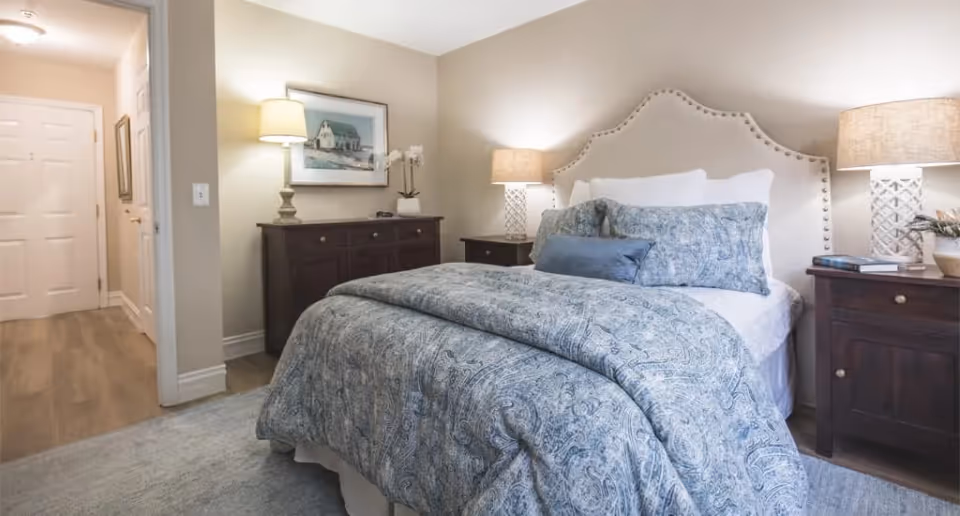 A cozy bedroom featuring a bed with a patterned blue and white comforter and multiple pillows. The bed has a beige upholstered headboard with nailhead trim. On either side of the bed are dark wooden nightstands with matching lamps. A dresser with a lamp and a framed picture is visible against the far wall. The room has light-colored walls and a carpeted floor, with a hallway leading to a white door.