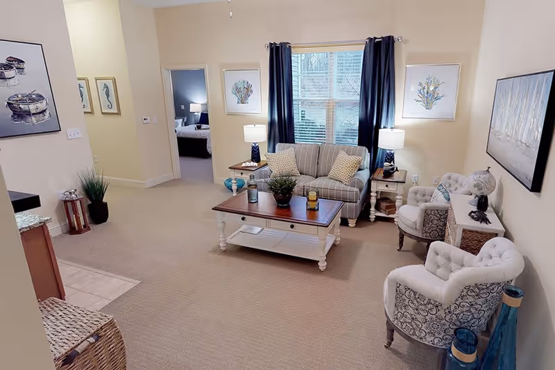 A cozy living room with a striped loveseat, two patterned armchairs, a wooden coffee table with decorative items, two side tables with lamps, and framed artwork on the walls. A window with dark curtains allows natural light into the room. A bedroom is visible through an open doorway.