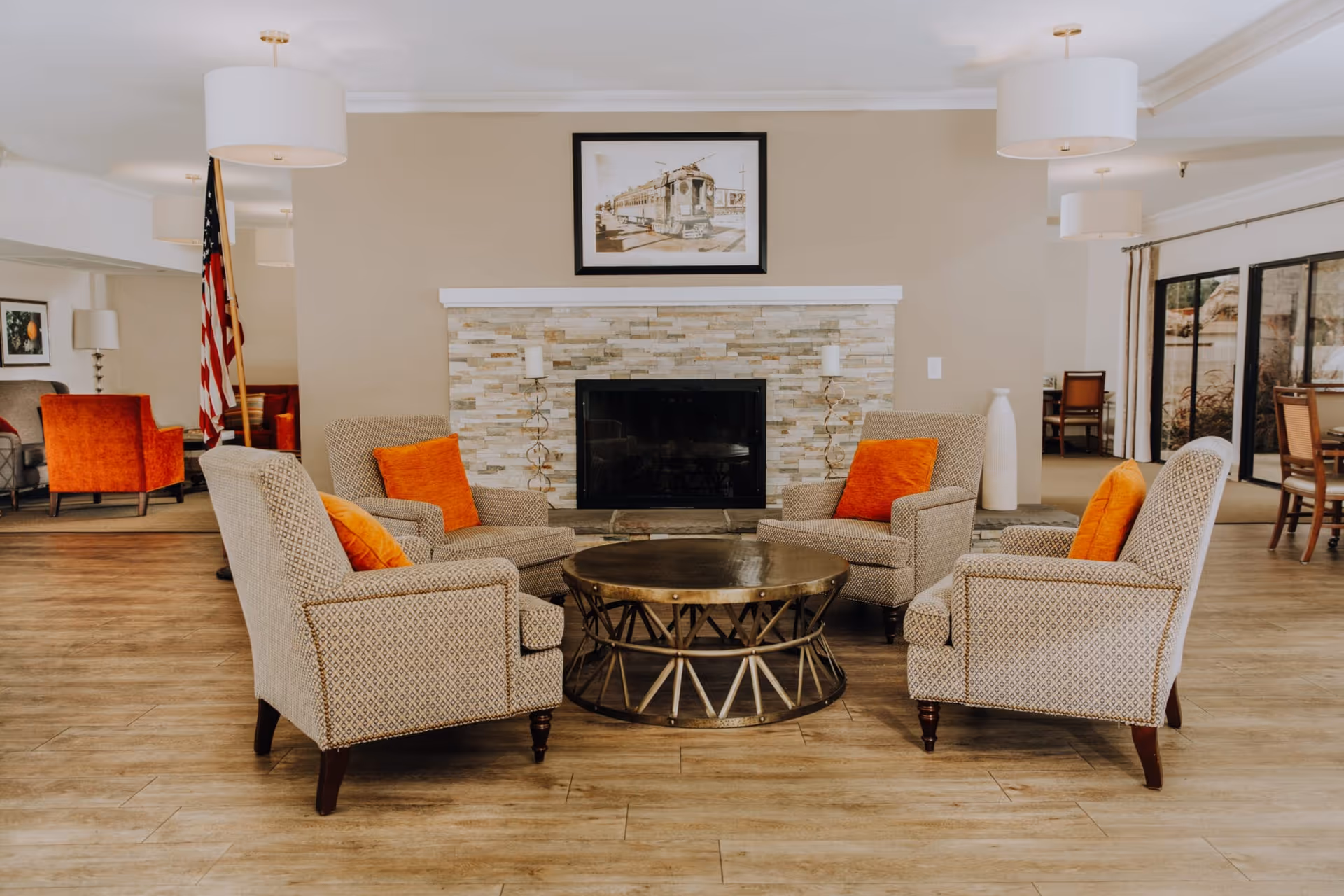 A cozy senior living common area with four patterned armchairs arranged around a round metal coffee table. Each chair has an orange cushion. The room features a stone fireplace with two candle holders on either side and a framed picture of a streetcar above it. The floor is wood, and there are large windows and additional seating visible in the background.