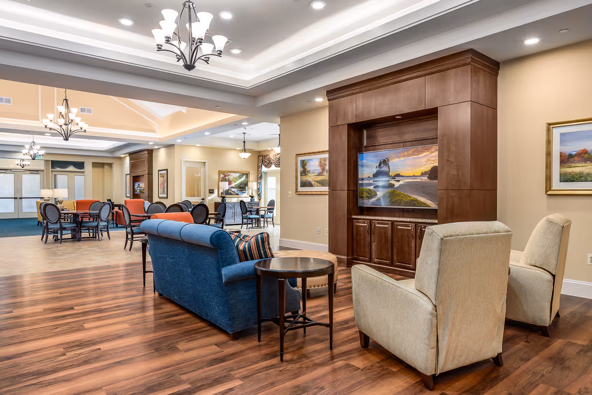 A spacious and well-lit common area in a senior living facility featuring a blue sofa, two beige armchairs, a round side table, and a large wooden entertainment center with a flat-screen TV displaying a scenic beach sunset. The room has wood flooring, beige walls adorned with framed paintings, and multiple dining tables with chairs in the background. Elegant chandeliers hang from the ceiling, adding to the warm and inviting atmosphere.