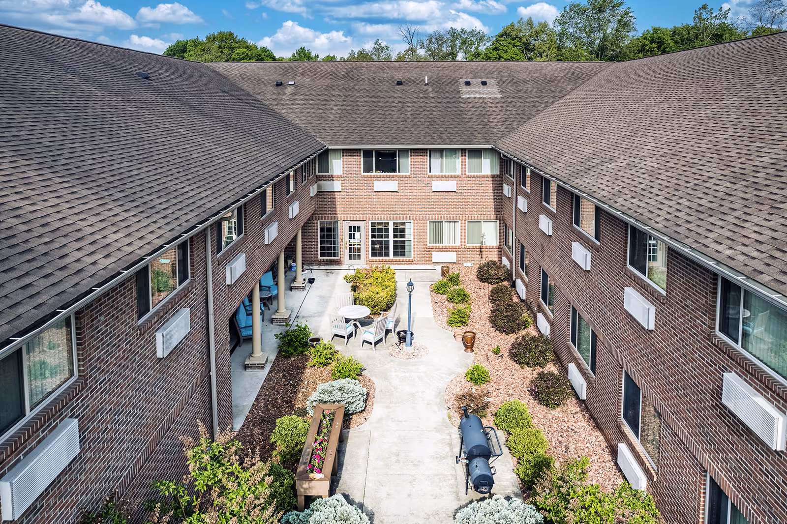 Aerial view of a brick senior living facility courtyard with a concrete walkway, outdoor seating area with table and chairs, landscaped garden beds with shrubs and flowers, and a barbecue grill. The building has two stories with multiple windows and a shingled roof under a partly cloudy sky.