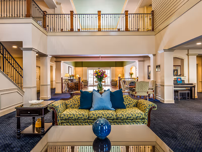 Open two-story lobby with a patterned sofa and pillows facing a coffee table, seating areas, flowers, and an upper balcony.
