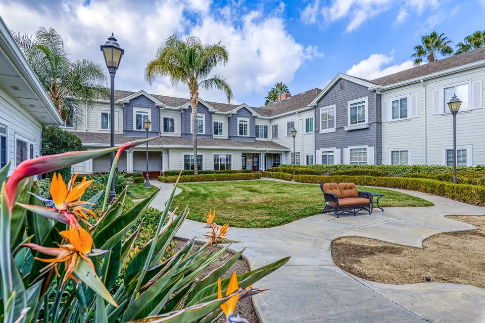 Outdoor courtyard area of a senior living facility with a well-maintained lawn, palm trees, flowering plants, a paved walkway, and a cushioned outdoor bench. The building surrounding the courtyard is two stories with white and gray siding and multiple windows under a partly cloudy sky.