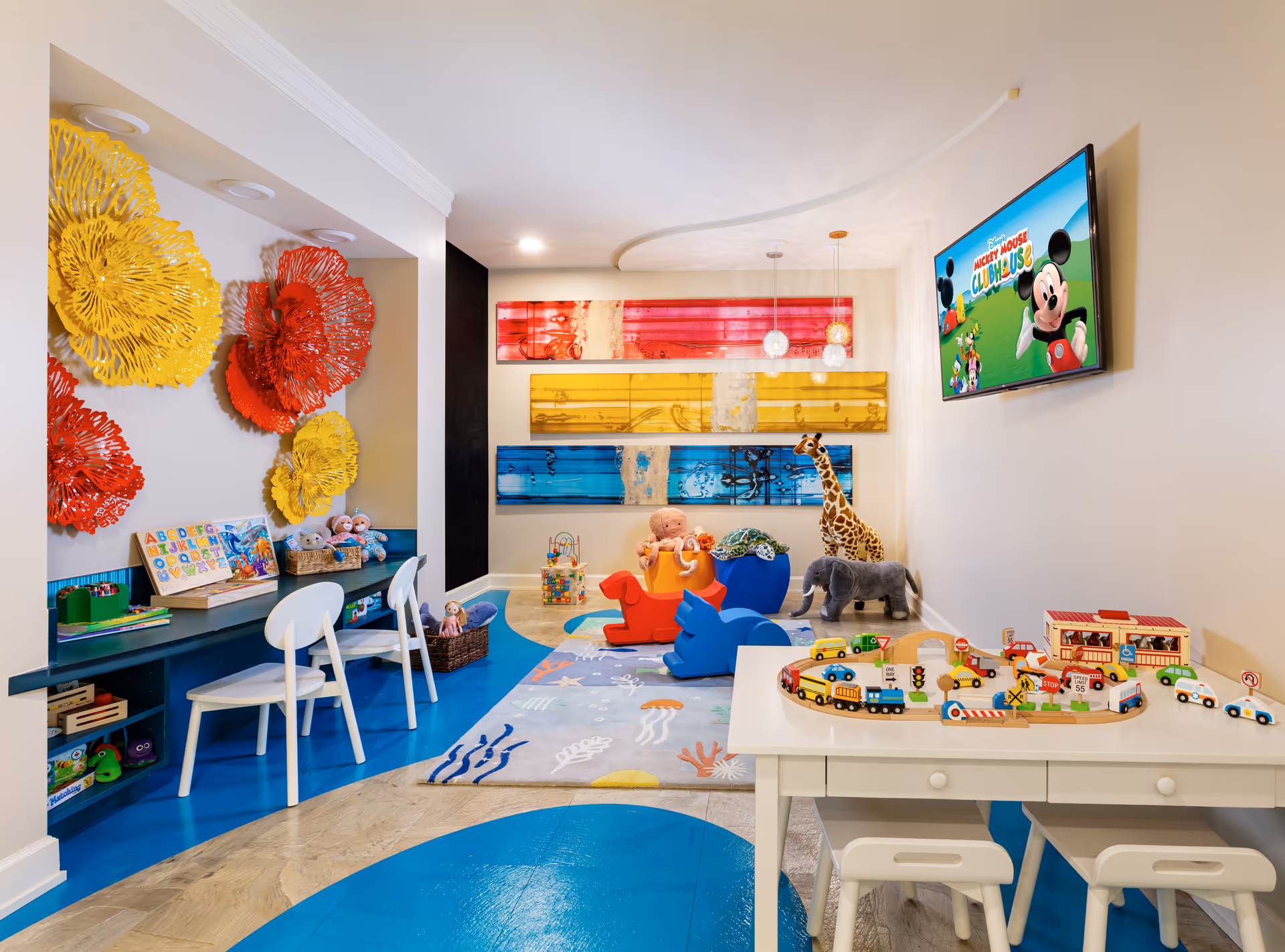A colorful children's playroom with a blue and beige floor, white walls, and vibrant wall decorations in yellow, red, and blue. The room features a small white table with stools and a wooden train set, a play area with toys including a giraffe, elephant, and rocking horse, and a wall-mounted TV displaying Mickey Mouse Clubhouse. There are also two white chairs at a built-in desk with books and toys.