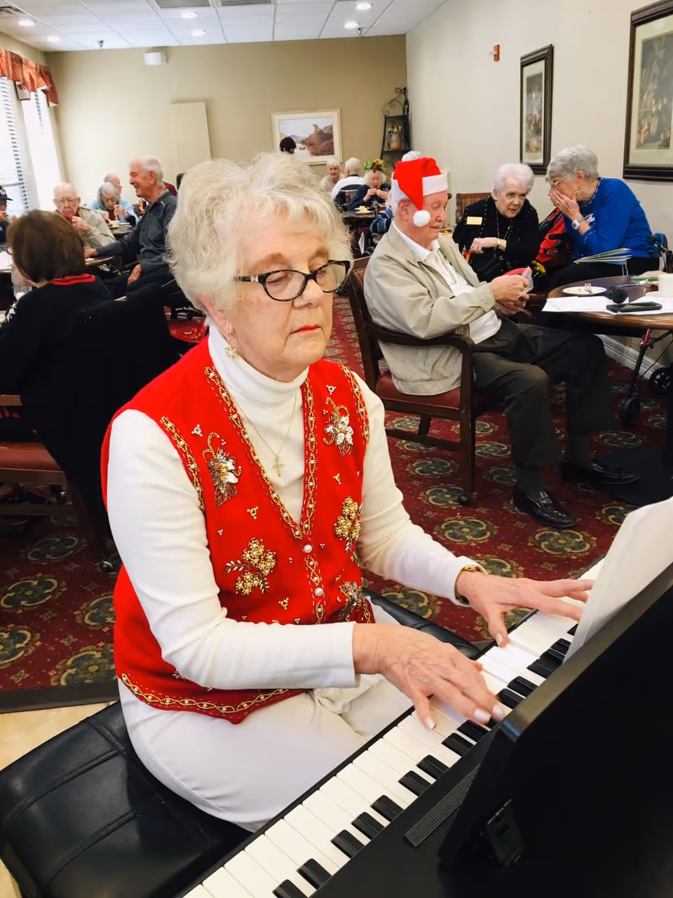 An elderly woman wearing glasses and a red festive vest is playing a keyboard piano in a room filled with other elderly people sitting at tables. One man in the background is wearing a Santa hat. The room has framed pictures on the walls and a patterned carpet.