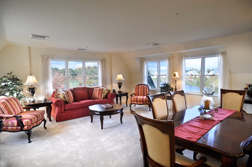 A bright and spacious living and dining area with large windows showing a scenic outdoor view. The living area features a red sofa, two striped armchairs, a wooden coffee table, and side tables with lamps. The dining area has a wooden table with six upholstered chairs and a red table runner with a decorative centerpiece.