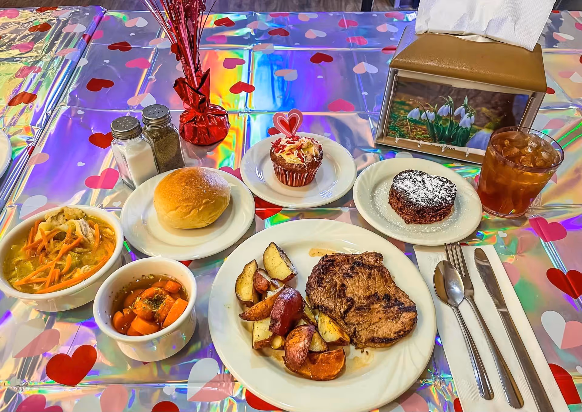 A meal served on a table with a shiny, heart-patterned tablecloth. The meal includes a bowl of soup with vegetables, a small bowl of cooked carrots, a bread roll on a plate, a plate with grilled meat and roasted potatoes, a heart-shaped chocolate dessert dusted with powdered sugar, a cupcake with a heart decoration, a glass of iced tea, salt and pepper shakers, and a napkin holder with a floral picture.