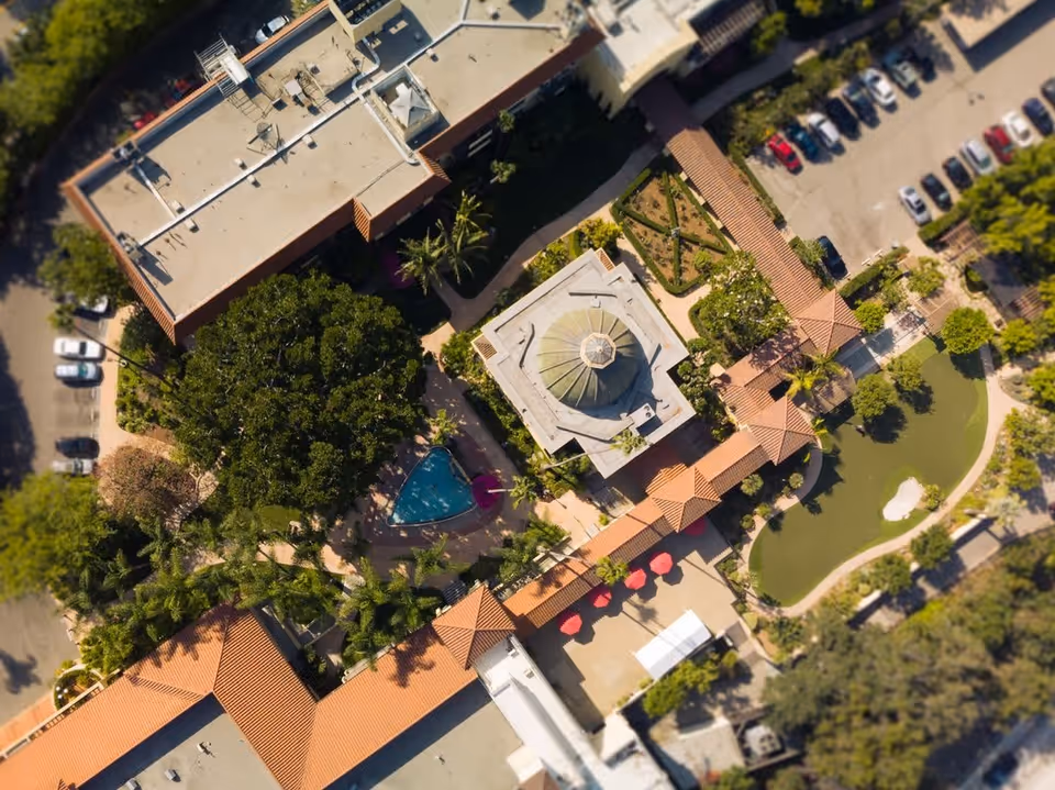 Aerial view of Hollenbeck Palms senior living facility showing buildings with red-tiled roofs, a small triangular swimming pool, landscaped gardens, walking paths, a putting green, and a parking lot with cars.