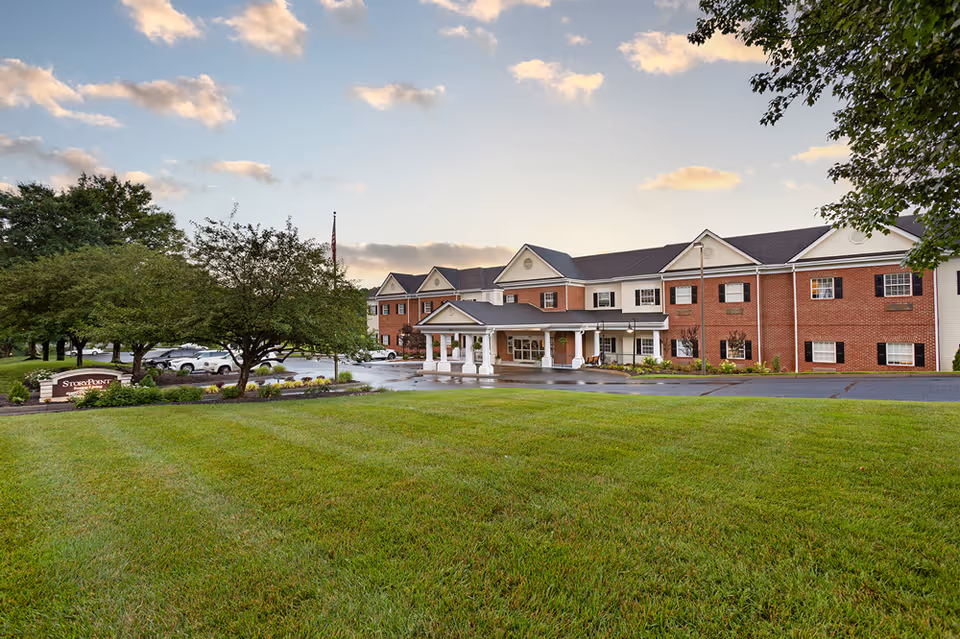 Exterior view of a two-story senior living facility building with a brick facade and white trim. The building has a covered entrance with columns and a driveway in front. There is a well-maintained green lawn in the foreground, trees on the left and right sides, and a partly cloudy sky at sunset. A sign near the trees reads 'StoryPoint'.