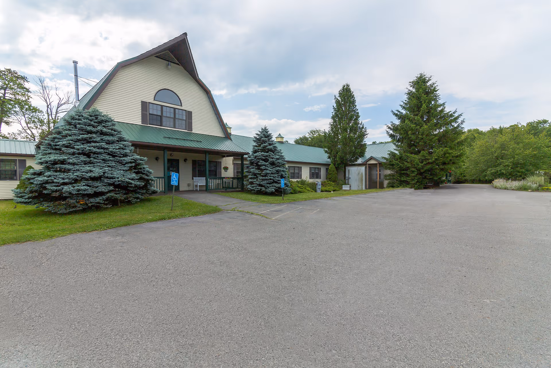 Front exterior of a senior living building with a green metal roof, large evergreen trees, and an empty paved parking area.
