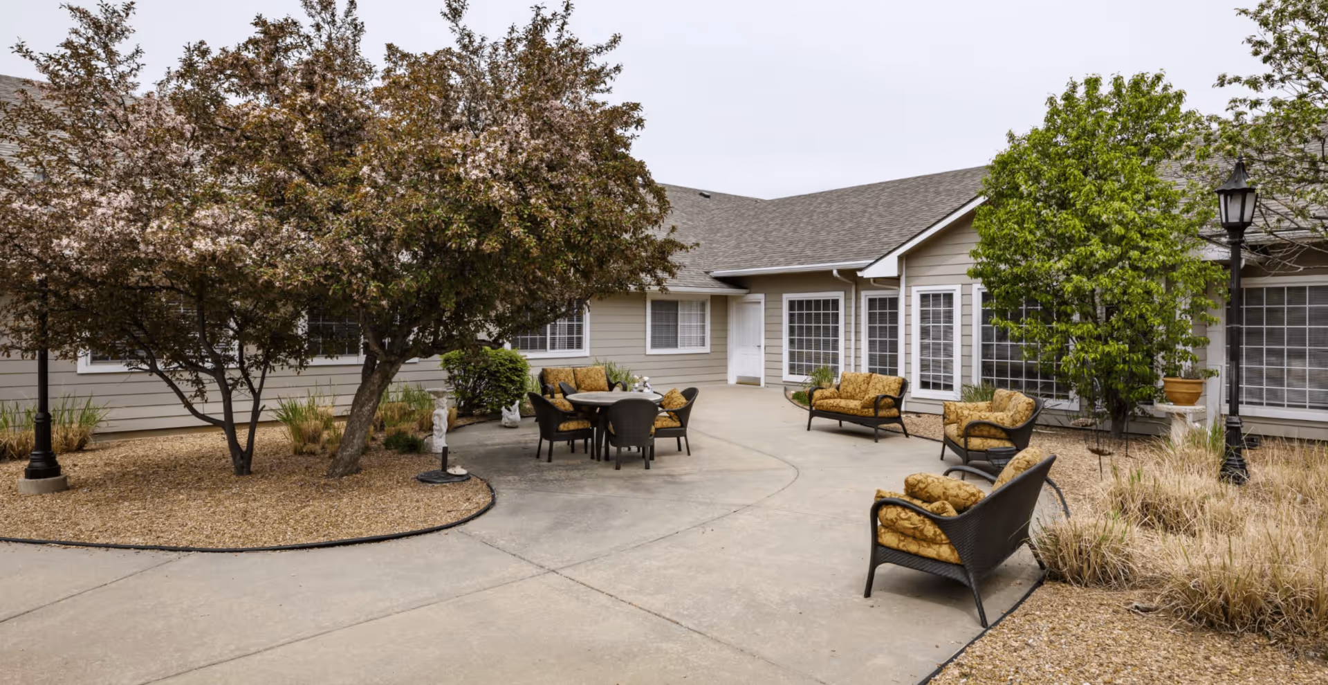 Outdoor courtyard area at Homestead Estates of Wichita featuring a paved walkway, several cushioned wicker chairs and a table, surrounded by trees and landscaping with bushes and ornamental grasses.