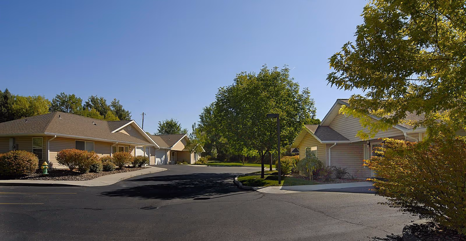 View of a senior living community street with single-story beige buildings on either side, surrounded by trees and bushes under a clear blue sky.