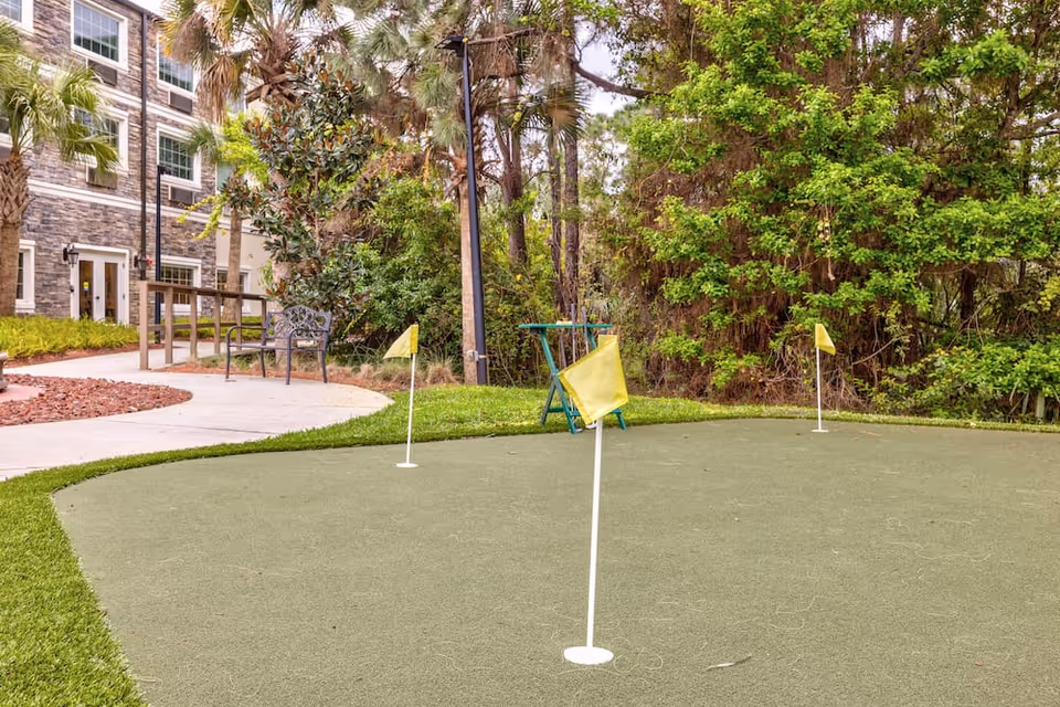 Small outdoor putting green with three yellow flags in front of a multi-story building and surrounding trees.