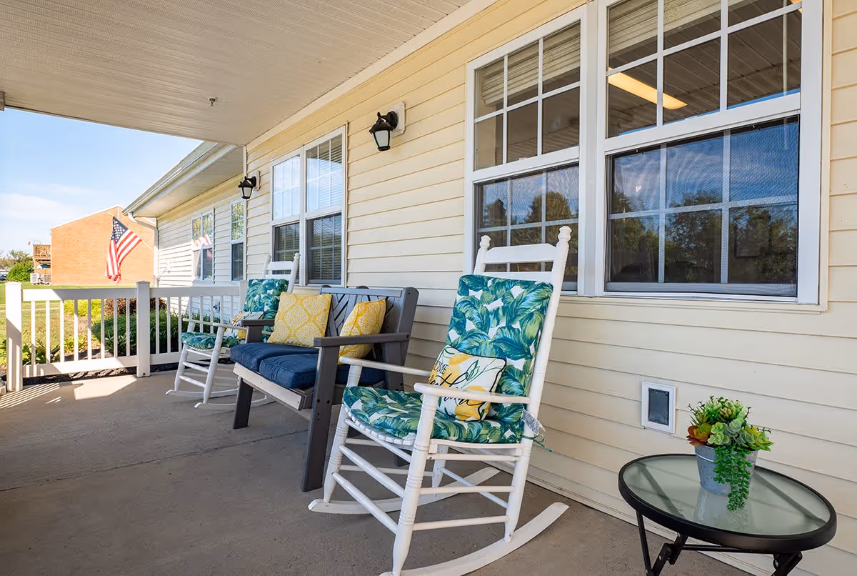 A covered outdoor porch area with white rocking chairs and a bench with colorful cushions. There is a small round glass table with a potted plant on it. The porch has beige siding and white framed windows, with an American flag visible in the background.