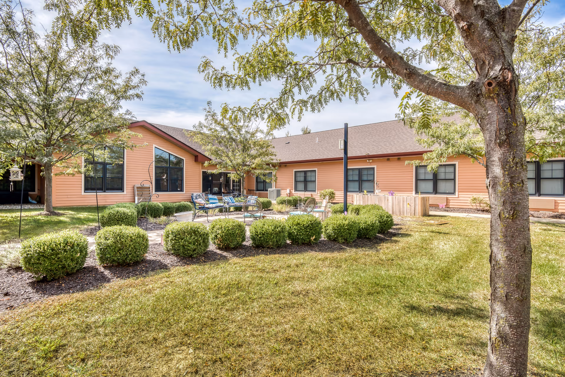 Outdoor garden area at The Meadowlands featuring a grassy lawn, neatly trimmed bushes, several trees, and a seating area with chairs and a table. The building in the background is a single-story structure with peach-colored siding and multiple windows.