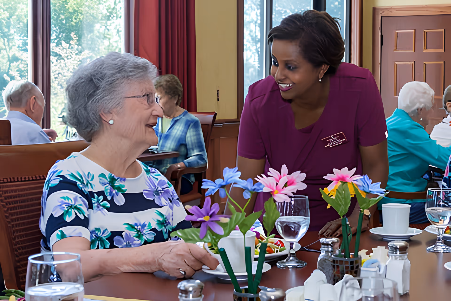 A caregiver leans in to chat with a smiling elderly woman seated at a table set with flowers and dishes in a senior dining room.