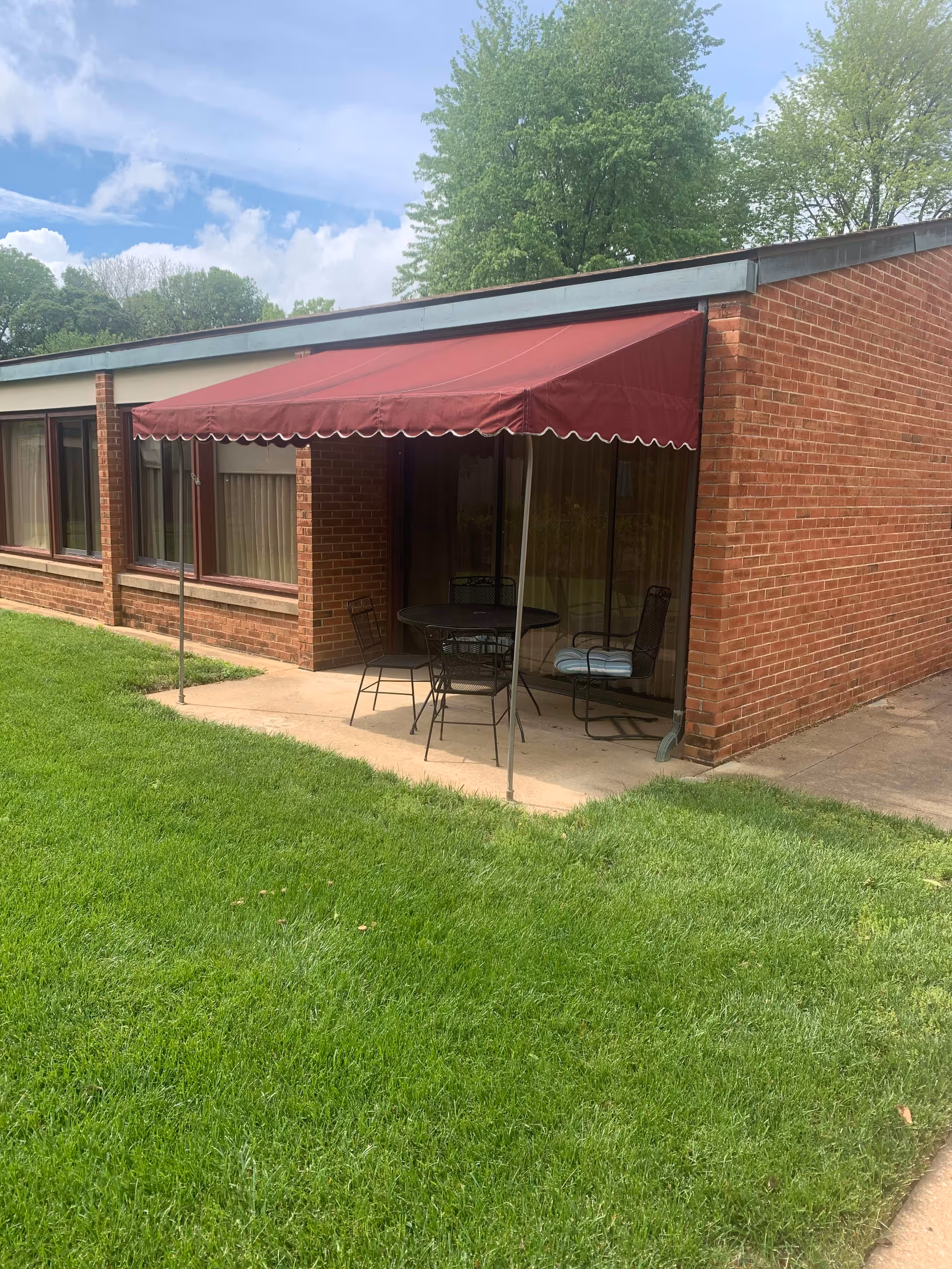 Outdoor patio area at a brick building with a maroon awning, a round table, and four metal chairs on a concrete slab surrounded by green grass and trees in the background under a partly cloudy sky.