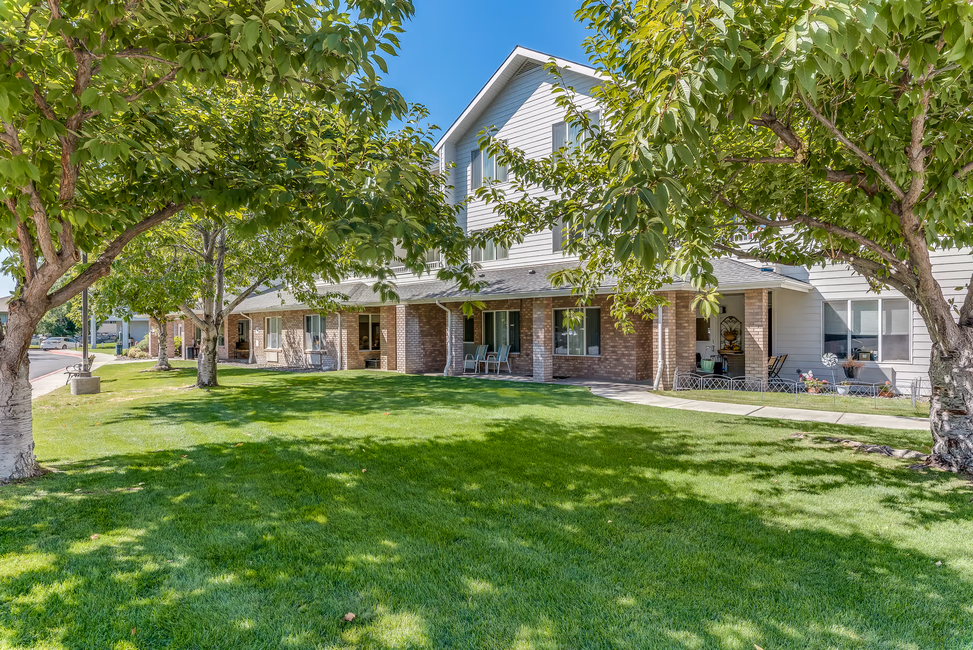 Front exterior of a two-story senior living building with a green lawn, trees, and covered porches with chairs.