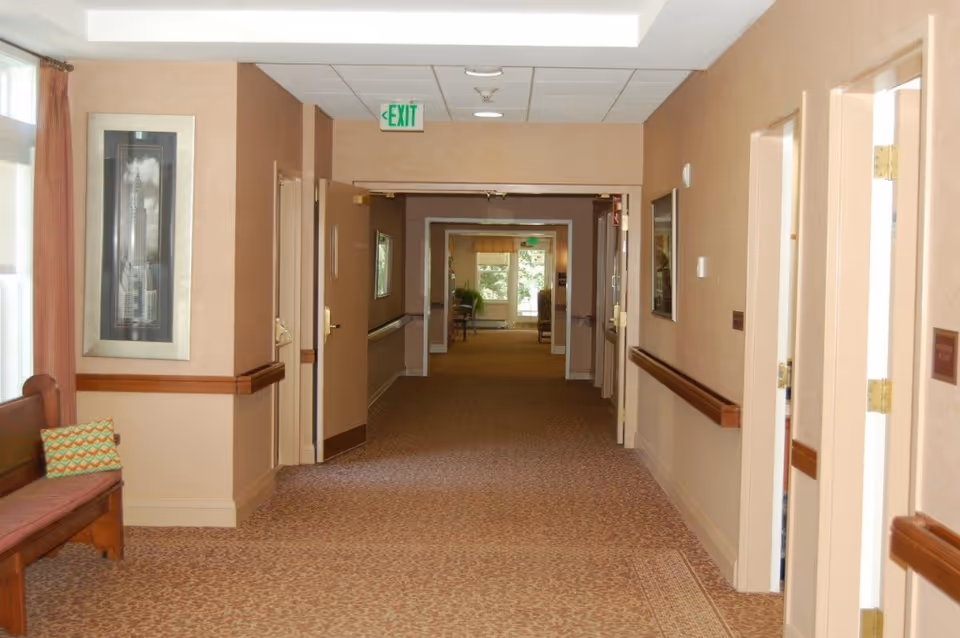 Carpeted interior hallway of an assisted living facility with handrails, doors, a bench, and an EXIT sign.