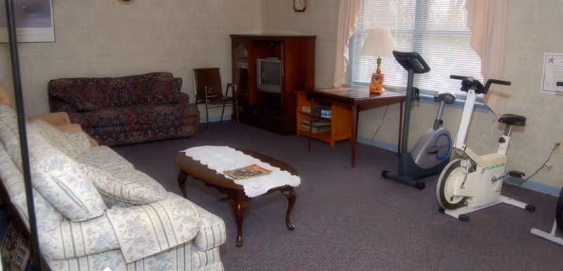 A living room with two patterned sofas, a wooden coffee table with a white lace runner and magazines, a wooden TV stand with a small television, a wooden side table with a lamp, and two exercise bikes near a window with pink curtains.