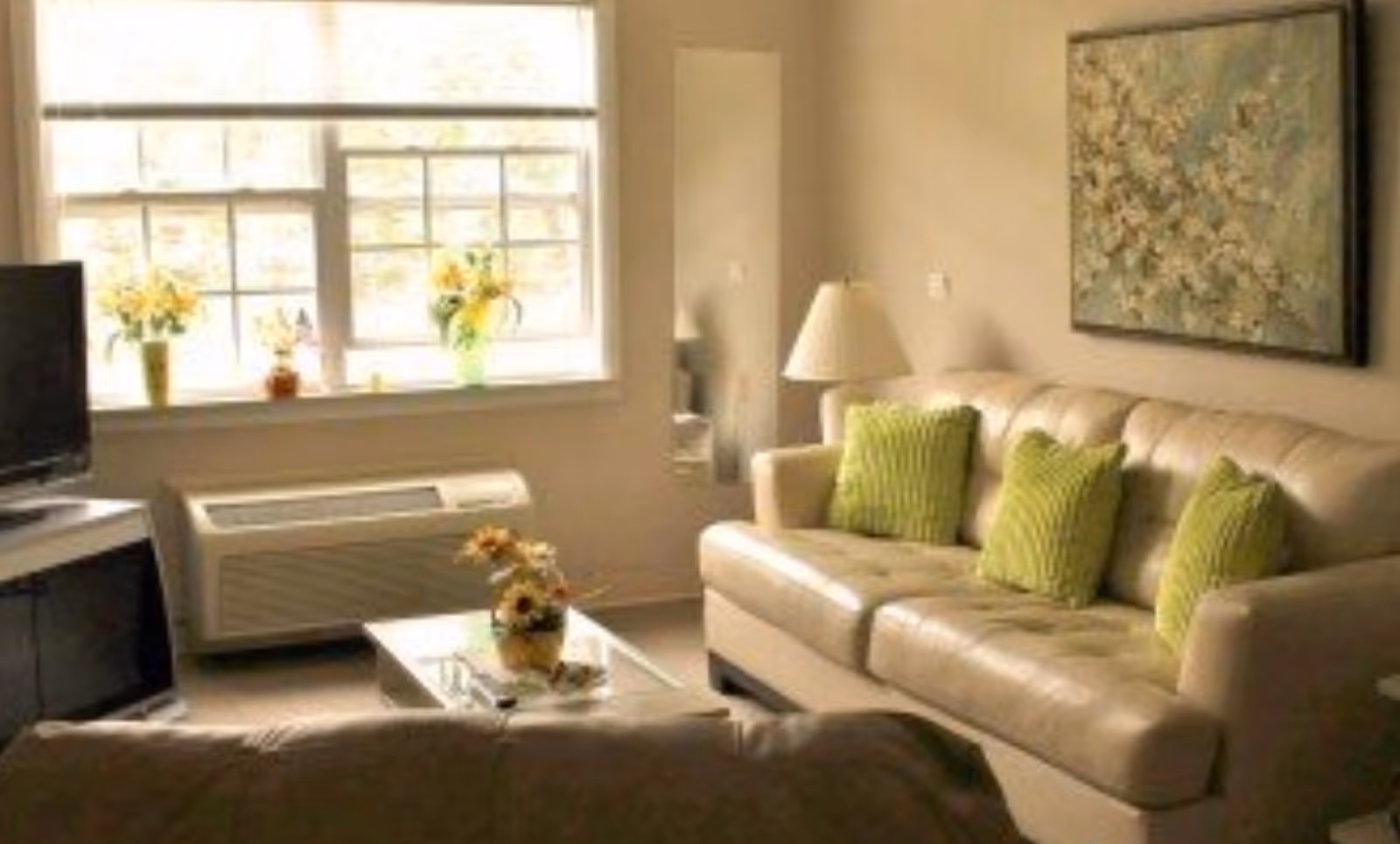 A cozy living room with a beige leather sofa adorned with three green pillows, a glass coffee table with a flower arrangement, a TV on a stand, a window with flower vases on the sill, a floor lamp, and a floral painting on the wall.